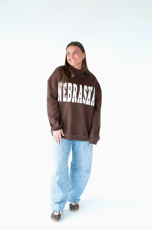 Model wearing a brown Nebraska mockneck sweatshirt with white collegiate lettering