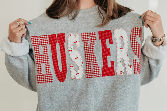 Model wearing a retro gray Nebraska Huskers crewneck sweatshirt with red gingham and white heart-pattern HUSKERS lettering.
