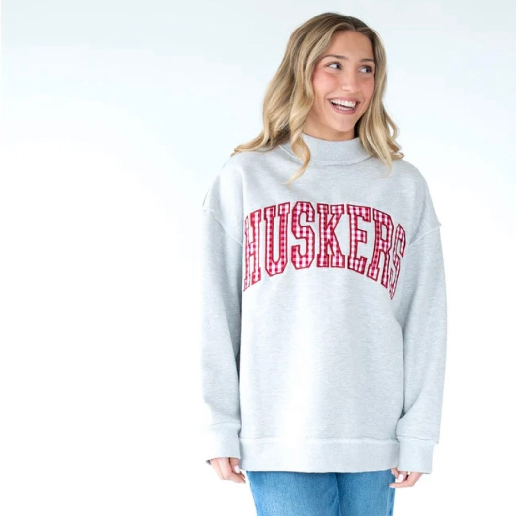 Woman wearing a heather gray Nebraska Huskers mockneck sweatshirt with red gingham arched HUSKERS lettering and an oversized fit.