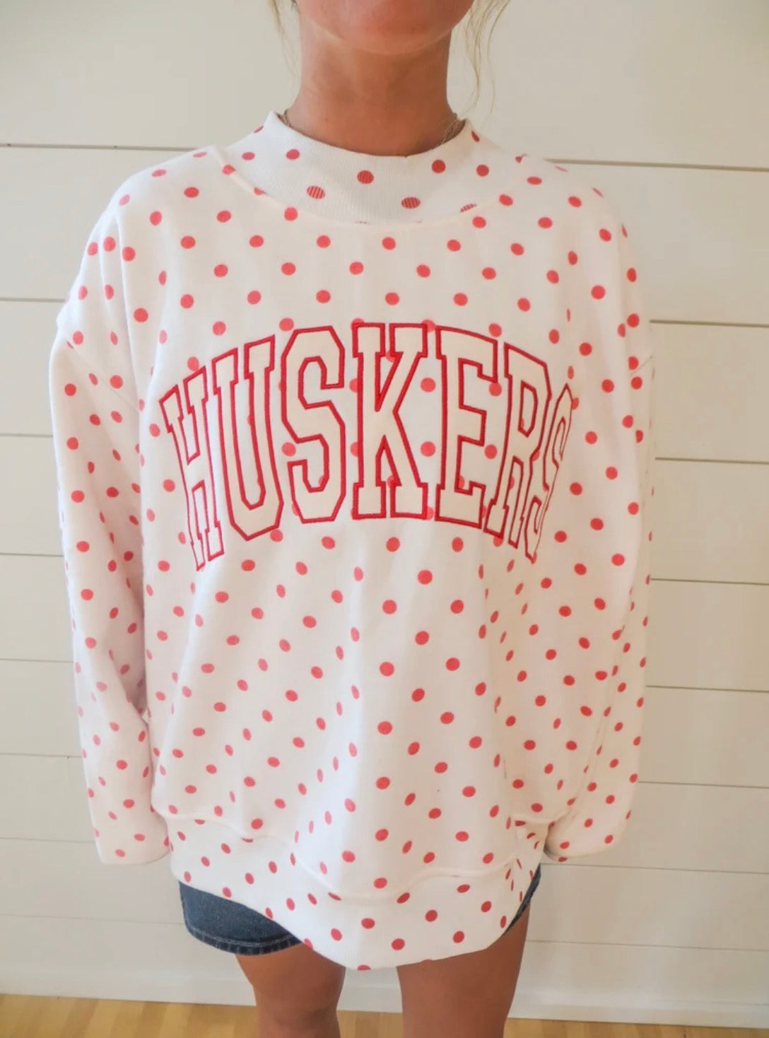 Close-up of a white Nebraska Huskers mockneck sweatshirt featuring red polka dots and outlined arched HUSKERS lettering.