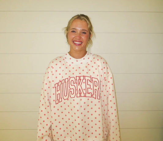 Woman wearing a white Nebraska Huskers mockneck sweatshirt with red polka dots and large arched HUSKERS lettering.