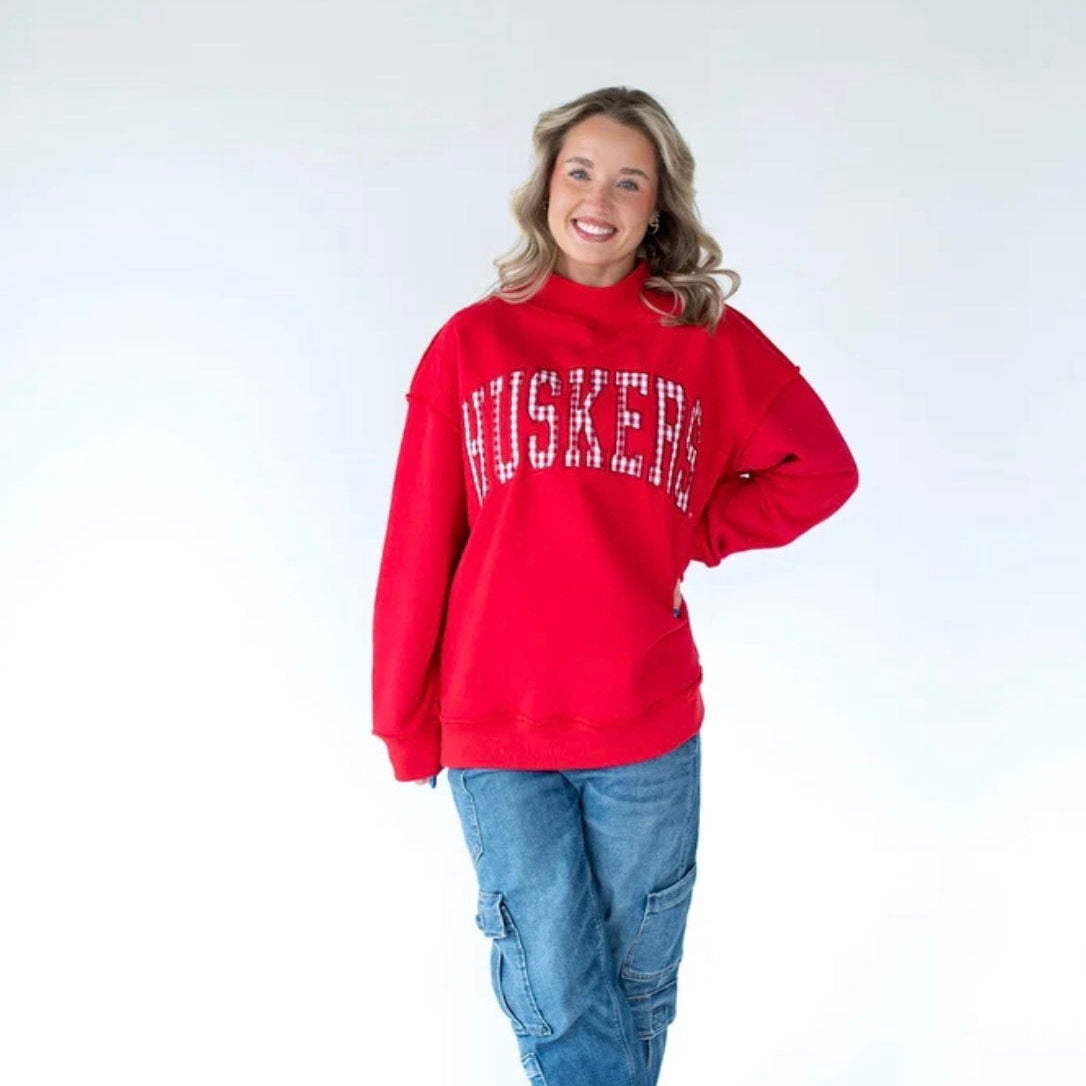 Woman wearing a Nebraska Huskers red mockneck sweatshirt with white gingham arched HUSKERS lettering and a relaxed oversized fit.