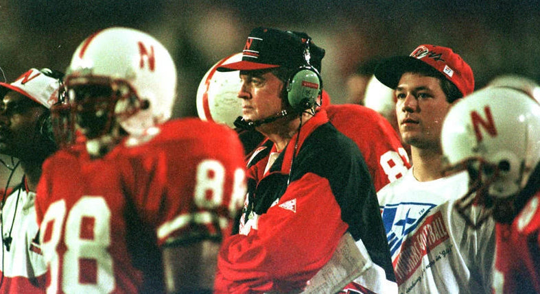 Vintage Nebraska Cornhuskers football sideline with players and Tom Osborne in red and white uniforms during a night game.