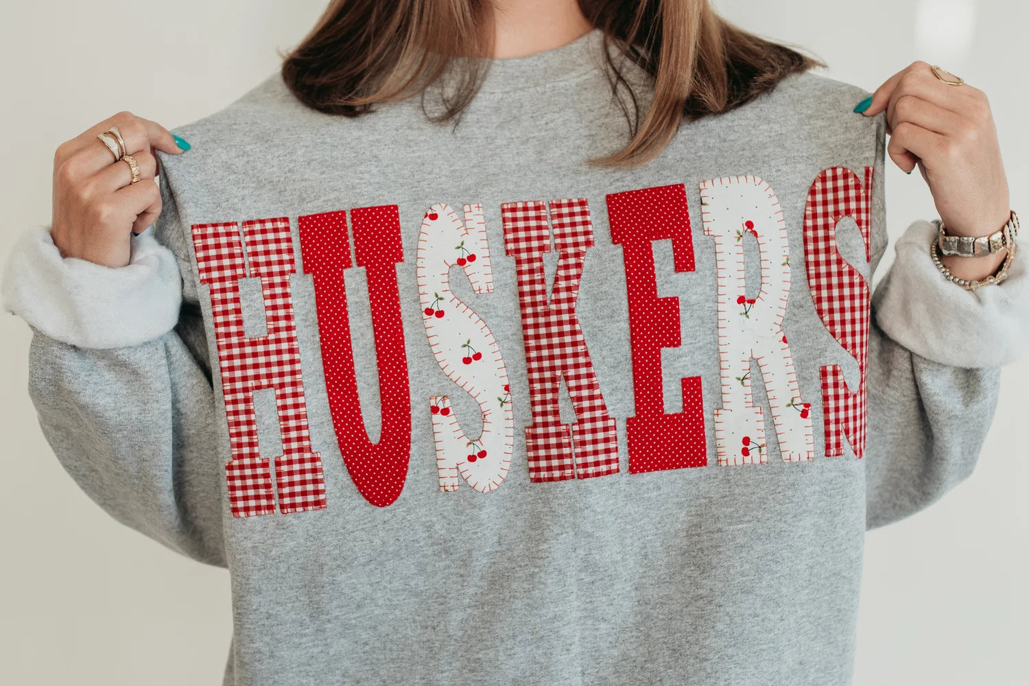Model wearing a retro gray Nebraska Huskers crewneck sweatshirt with red gingham and white heart-pattern HUSKERS lettering.