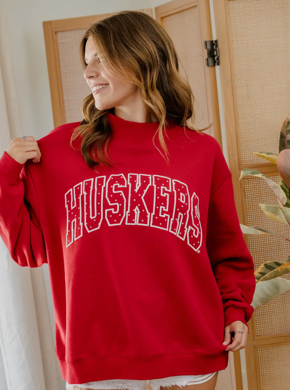 Side view of model wearing a red Nebraska Huskers mockneck sweatshirt with white polka dot HUSKERS graphic.